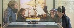 A group of children look intently at the info screen for a Sail boat housed in a glass case.
