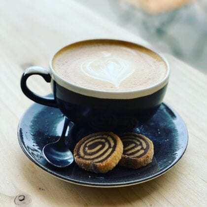 A latte with a heart in a large black mug on a saucer, coupled by 2 small biscuits.