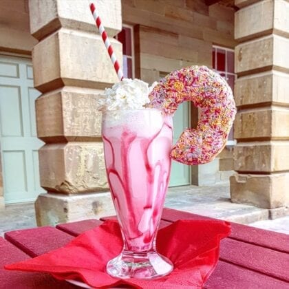 A pink milkshake, decorated by a large, pink, sprinkled doughnut and whipped cream.
